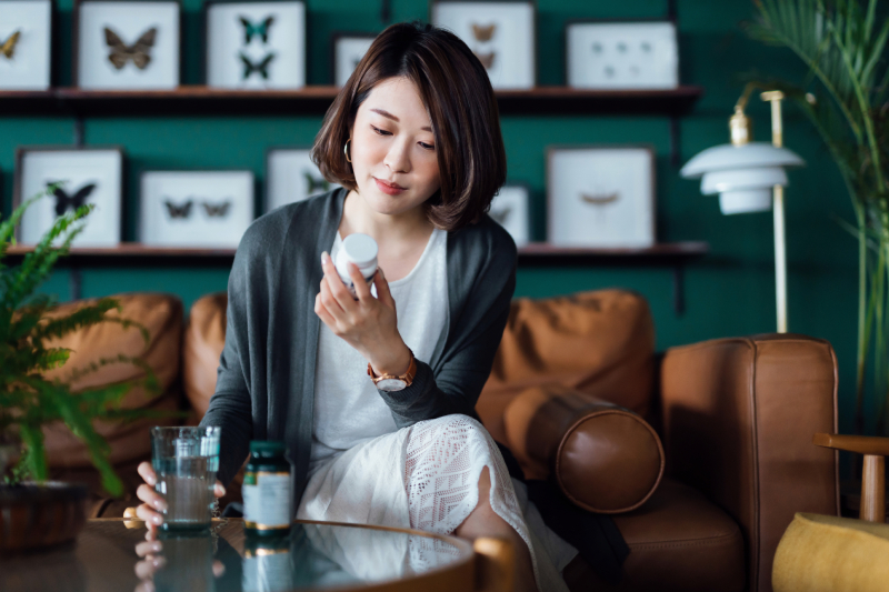 Young Asian woman taking medicine with a glass of water on the coffee table, reading the information on the label of her medication.