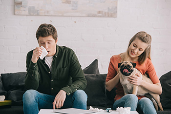 man, woman and pug dog sitting on couch with man holding a tissue to his nose